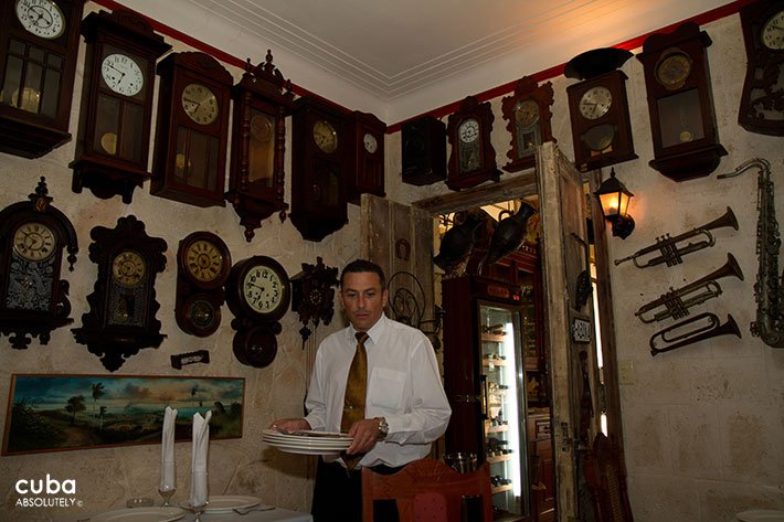 Waiter carrying dishes ina restaurant with a wall full of differents clocks © Cuba Absolutely, 2014